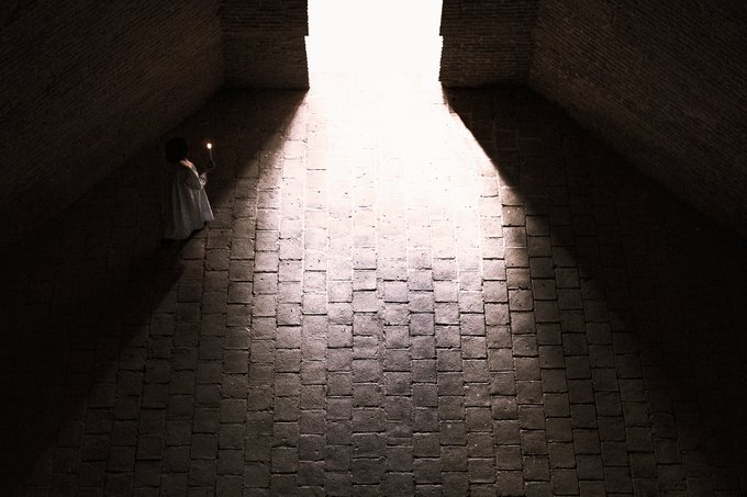 A dimly lit brick passageway opens to a bright white exit, where a small figure stands holding a light near the shadowed left wall.