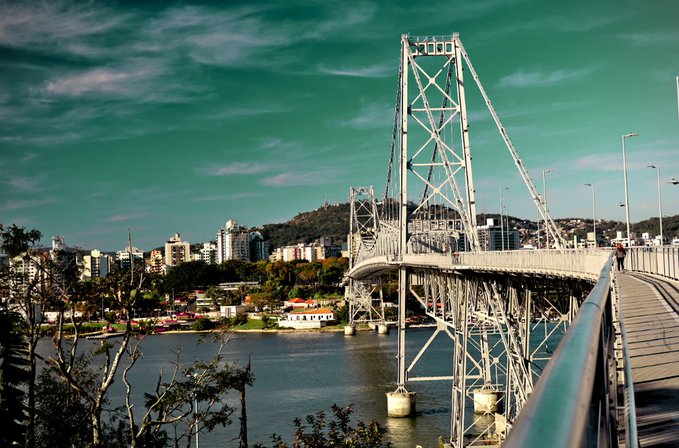 A dramatic teal-and-amber view of a steel suspension bridge over a city waterfront, with hills, buildings, and boats in the background. The altered colors reinforce the post’s theme of artistic freedom—“Art begins when you stop using the colors they chose for you”—while the visible link directs viewers to the artwork on objkt.com.