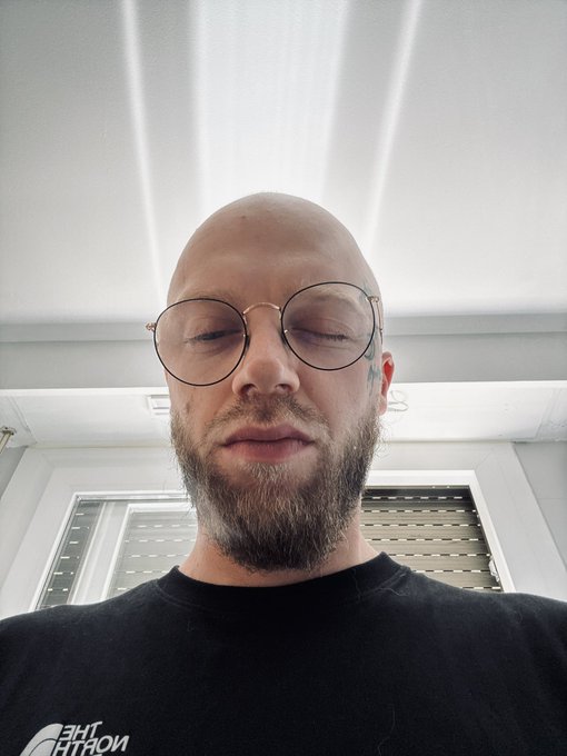 A bald man with a beard and round glasses looks downward at the camera, wearing a black "The North Face" t-shirt, captured in a low-angle selfie inside a room with white walls and a window. The caption "gm getting caffeinated now welcome to the crypto life" suggests a morning coffee ritual tied to the crypto community's common "gm" (good morning) greeting.
