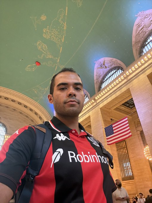 A man wearing a black and red Robinhood App soccer jersey takes a selfie inside what appears to be Grand Central Terminal, with the iconic green zodiac ceiling and American flag visible in the background. The image reflects his post as he sports Robinhood gear while representing his American identity and involvement in the crypto/Web3 space.