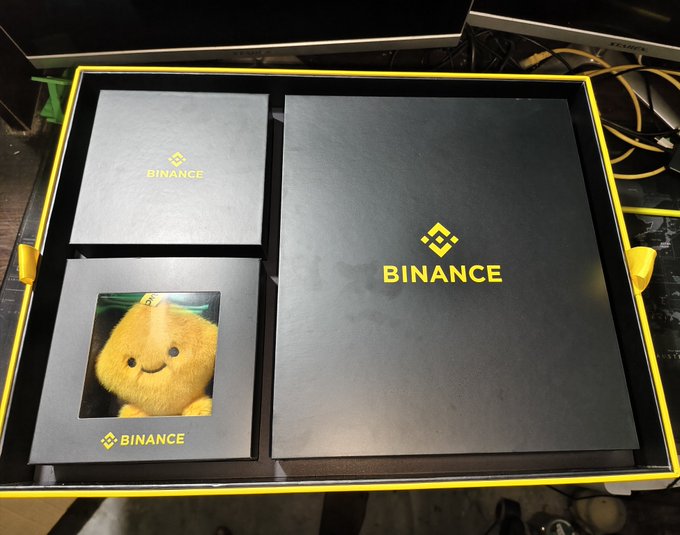 A black and yellow Binance promotional box containing branded envelopes and a card featuring a yellow smiling plush toy.