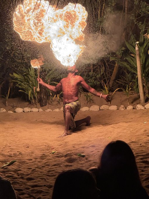A performer kneels on sandy ground, blowing a massive plume of fire from his mouth while holding a lit torch, during what appears to be a traditional fire-dancing show at night. The image doesn't directly relate to the post's discussion of SpacX's unlock event or xAI's integration into SpaceX, but its dramatic spectacle of controlled fire serves as a vivid metaphor for the explosive financial and corporate developments being analyzed.