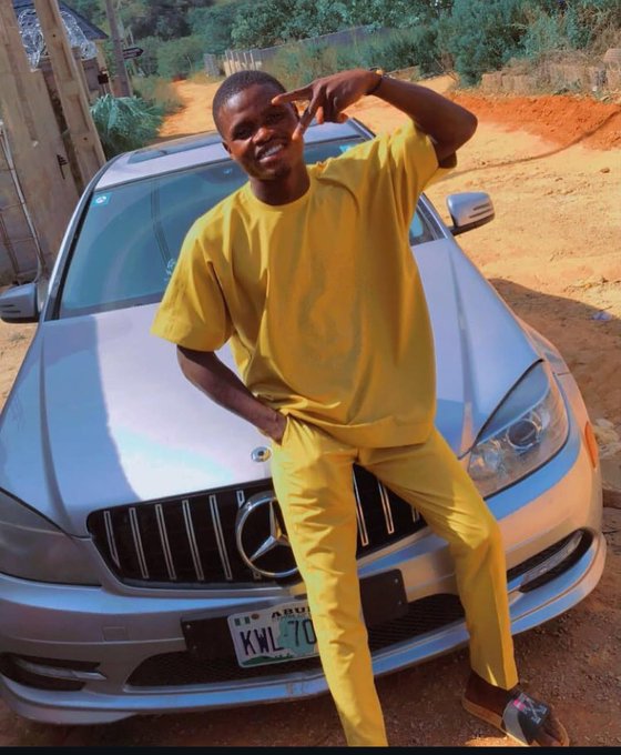 A smiling young man wearing a yellow shirt and pants poses with a peace sign in front of a silver Mercedes-Benz on a dirt road, with a license plate reading 'KWL-1C'.