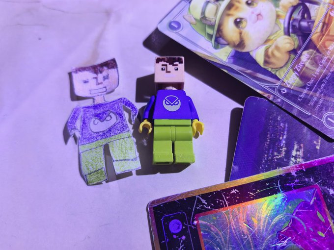 The image features a Lego minifigure standing next to another Lego minifigure with a drawing of a cat on a card, as well as some cards scattered around them. The scene appears to be set up for a play session or display, possibly related to a Minecraft-inspired project. The post includes various elements such as the Lego minifigures and cards, which are likely part of an imaginative and creative project involving both toys and cards.