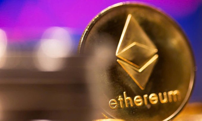 The image features a close-up of a gold coin with the word "Ethereum" written on it. The coin has a unique design that resembles an upside-down triangle, which adds visual interest to the piece. The background of the image is blurred, drawing attention to the coin and making it the focal point of the photo.