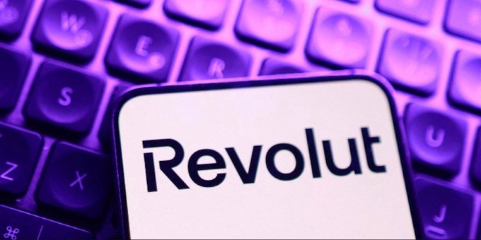 A close-up image shows the Revolut logo and name on a white smartphone screen, set against a backdrop of a purple-lit computer keyboard.