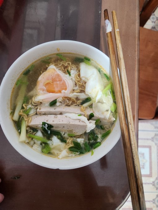 A bowl of noodle soup with a soft-boiled egg, sliced meat, and green vegetables in broth, served with wooden chopsticks on a dark wooden table.