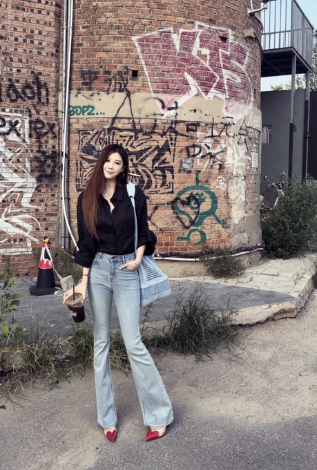 A young woman with long dark hair poses in front of a graffiti-covered brick wall, wearing a black blouse, light blue flared jeans, red pointed heels, and carrying a striped shoulder bag.