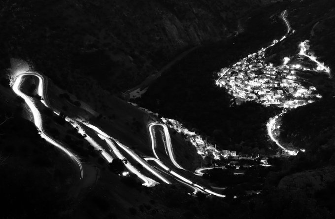 A black and white night-time aerial view showing the bright, winding light trails of a mountain road alongside an illuminated hillside village.