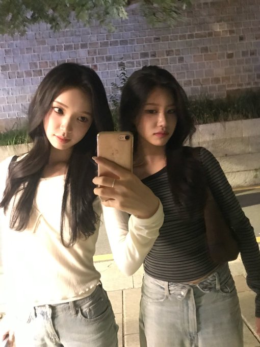 Two young Asian women taking a mirror selfie with a smartphone, one wearing a cream sweater and the other in a striped gray top with jeans.