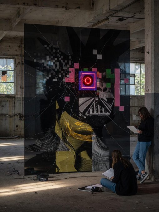 Two people examine a large, abstract black digital art installation with colorful geometric elements in an industrial space, with one person standing and another sitting on the floor with a notebook.