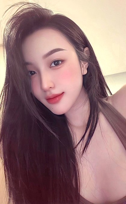 A selfie of a young woman with long dark hair, fair skin, and pink blush makeup in what appears to be an indoor setting.