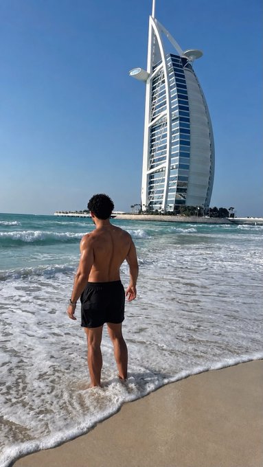 A shirtless man in black shorts stands at the water's edge on a sandy beach, gazing out at the iconic Burj Al Arab hotel in Dubai, the perfect backdrop for a crypto influencer boasting about massive gains on tokens like $ROSIE and $STAKE.