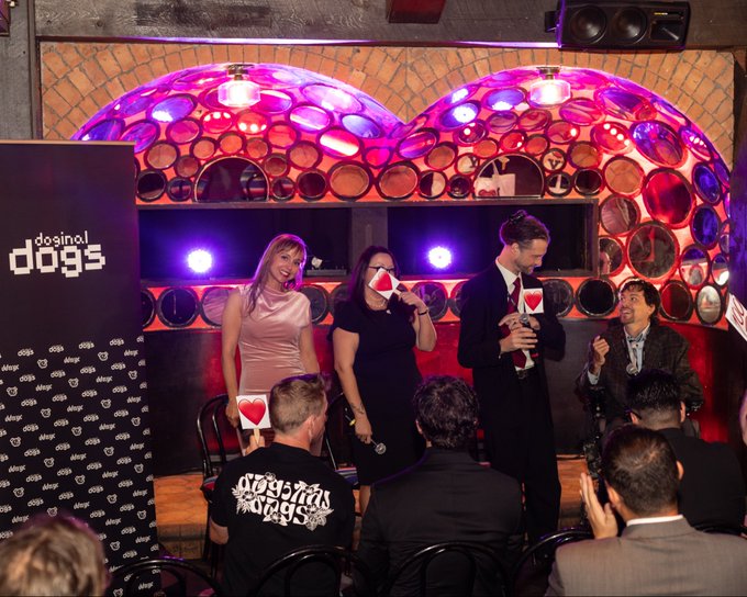 An indoor event scene with purple lighting and circular wall decor, showing several people including some wearing 'doginal dogs' branded clothing, with one person speaking into a mic while others hold cards with hearts on them.