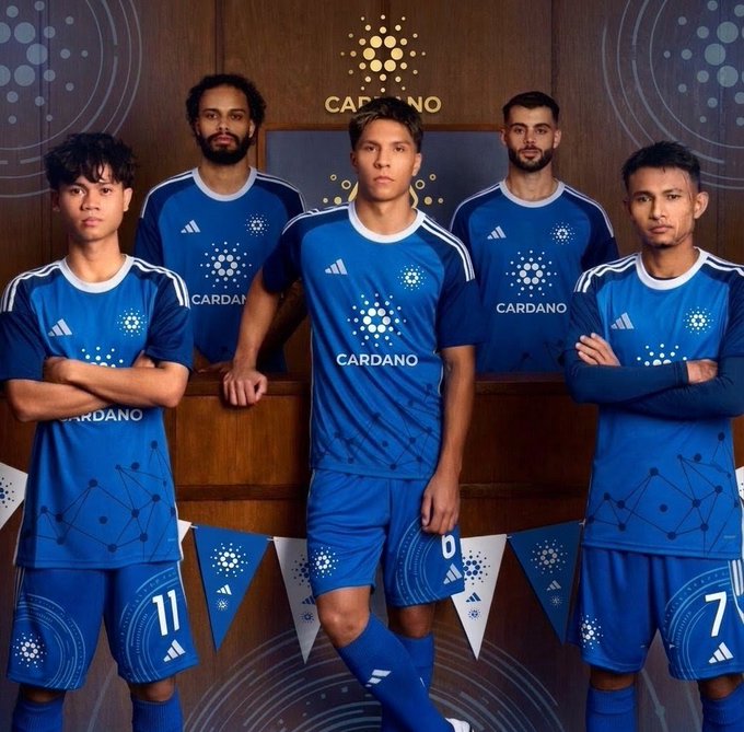 A group photo of five soccer/football players wearing blue Cardano-sponsored Adidas jerseys with the Cardano logo and blockchain-inspired design, standing in front of a Cardano-branded backdrop with pennant banners.