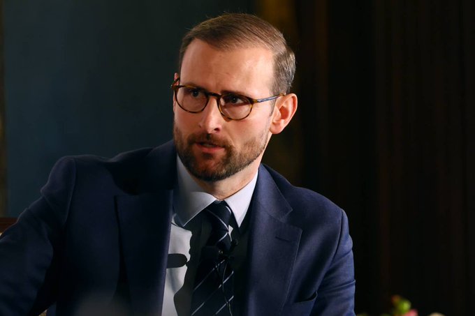 The image features a man wearing glasses, a suit, and a tie sitting at a desk with his hands resting on the table. The man appears to be looking off into the distance or deep in thought. The caption reads "LATEST- White House crypto advisor Patrick Witt says #Altseason #xrp #btc". This suggests that the image might have been taken during an event related to cryptocurrency, such as a conference or panel discussion at the White House.