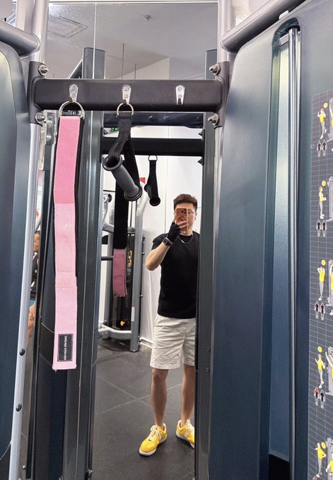 A mirror selfie taken inside a gym by a man wearing sunglasses, a black t-shirt, white shorts, and yellow sneakers, with pink resistance bands hanging from a cable machine.