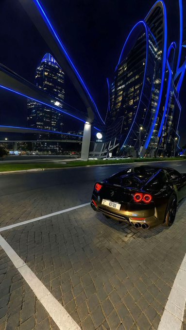 A nighttime photo of a gold/bronze sports car (with license plate '85 333') parked in front of a modern building illuminated with blue neon light streaks.