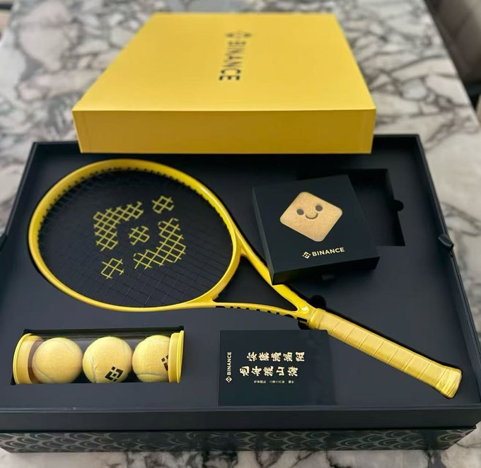 The image features a tennis racket with yellow and black colors, along with several tennis balls inside a box. The tennis racket has a handle that appears to be made of metal or wood. There are also two small cards placed next to the tennis racket in the box. The text "币安周边" is visible on one of the cards, which could potentially be related to the brand or model of the tennis racket and balls.

The main focus of the image is the tennis equipment displayed together, with a sense of sportyness and readiness for use in playing tennis.