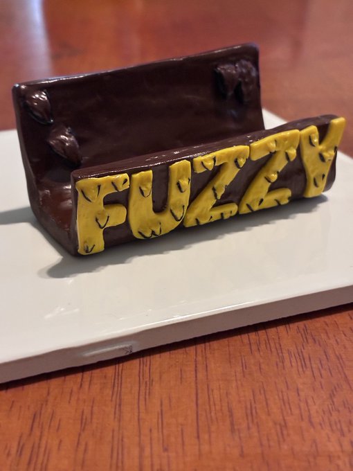 A hand-crafted dark brown clay card holder featuring the yellow lettering "FUZZY" sits on a white plate atop a wooden surface, serving as a handmade prototype being auctioned off to fund exploration of the DeepTide_xrp platform by the artist Metaphysic Clay.