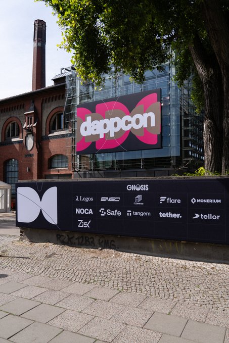 The image shows a large billboard with the text "Dappcon" written on it. The billboard also features logos for various companies such as "NOCA", "Zeiss", and "Ginnis". The backdrop of the billboard is a brick building, which adds an urban feel to the scene. In addition to these elements, there are trees visible in the background, providing some greenery to the image. The post about Dappcon seems to be promoting a conference or event related to data, computing, and finance.