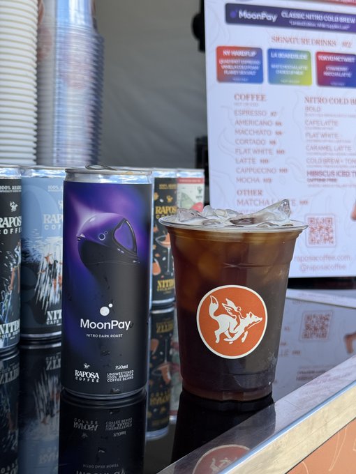 A can of MoonPay Nitro Dark Roast by Raposa Coffee sits next to an iced coffee in a branded cup, with a Raposa Coffee menu board listing nitro cold brews and signature drinks in the background.