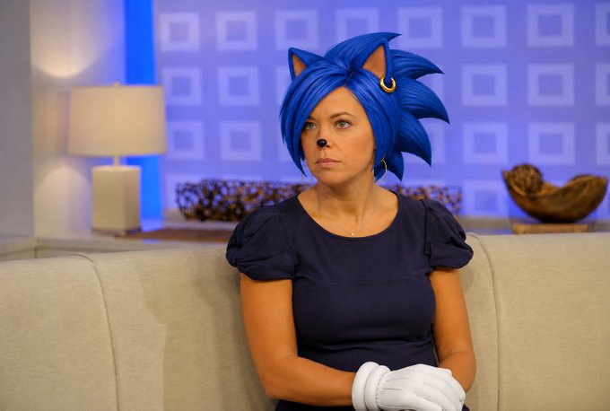 A woman dressed in a Sonic the Hedgehog costume—complete with a blue spiky wig, cat-like ears, and white gloves—sits on a beige couch in what appears to be a TV studio setting. The costume matches the "$sanic" reference in the post, a playful misspelling of the famous Sega character, tying the visual humor to the accompanying crypto-related tags.