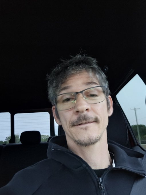 A middle-aged man with graying hair, a goatee, and glasses takes a selfie from inside a vehicle, wearing a black zip-up jacket.