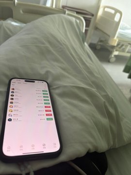 The image features a cell phone lying face down on top of a white pillow, which is placed on a bed. The phone screen displays an app with a list of names and numbers. The background shows a hospital room with beds and medical equipment visible in the distance.