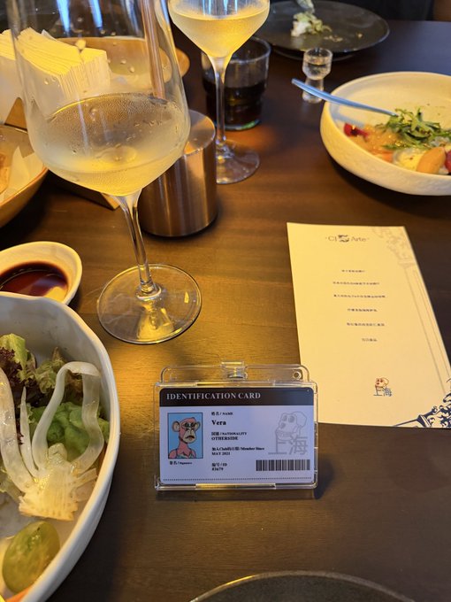 A restaurant table setting with wine glasses, dishes, and an identification card for "Vera" (Bored Ape Yacht Club #7100) displayed in a plastic holder, alongside a menu with Japanese text. The image ties into the "Join the club, Buy an ape" post by showing the practical use case of BAYC NFTs as membership-style identification cards at restaurants.