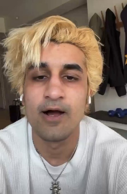 A young man with bleached blonde hair wearing a white sweater and a silver pendant necklace, appearing to be recording a video or speaking to camera.