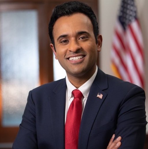 A professionally-dressed man, appearing to be Vivek Ramaswamy based on the post context, smiles confidently with arms crossed in a formal portrait setting, wearing a navy blue suit, red tie, and American flag lapel pin with a U.S. flag visible behind him. The image relates to the announcement of his company "Strive" purchasing $275 million worth of Bitcoin.