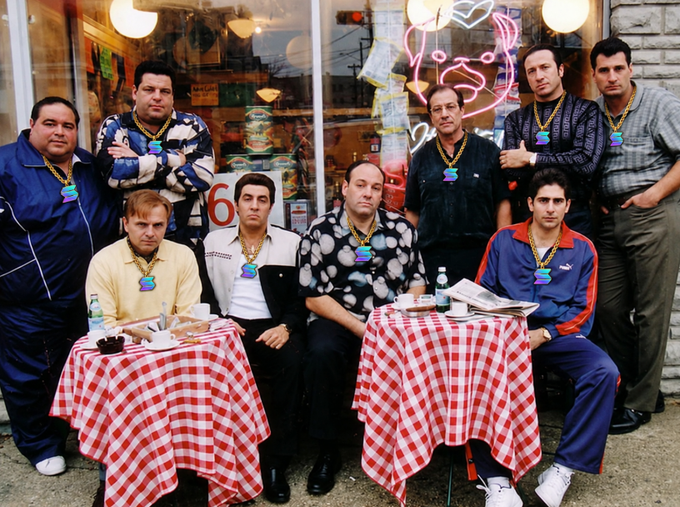 A group of nine men posing for a photo at an outdoor restaurant table covered with a red and white checkered tablecloth; they are wearing beaded necklaces with green pendants, and a neon sign and an Italian flag are visible in the shop window behind them.