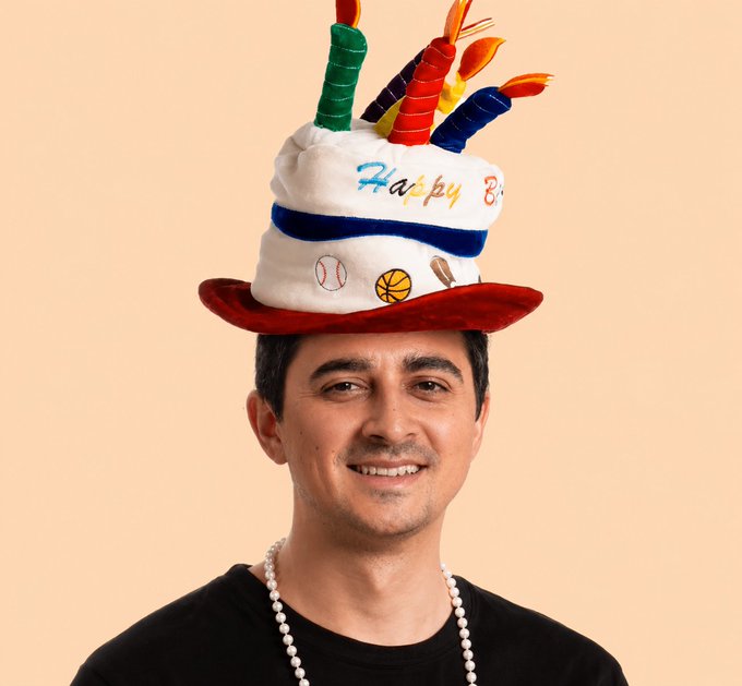 The image features a man wearing a birthday hat with candles on top, smiling for the camera. There are also several small objects scattered around him, including baseballs, a basketball, and a necklace. The background of the photo appears to be a light peach color, which complements the festive atmosphere created by the birthday hat.