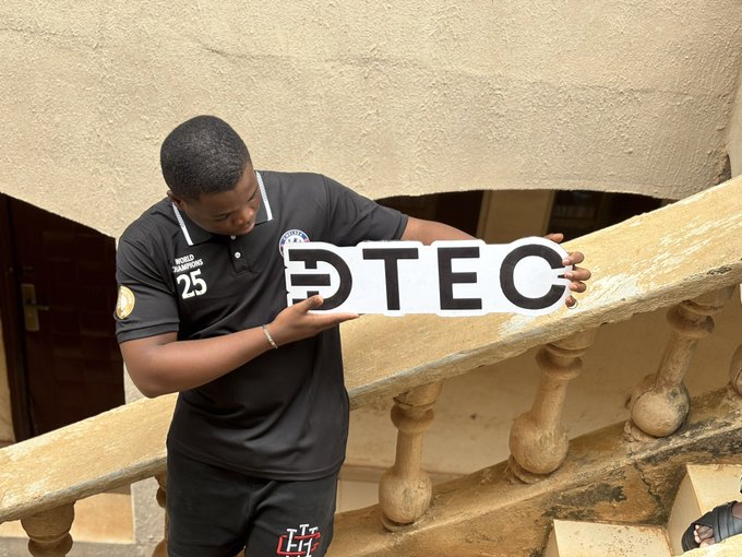 A young man wearing a black 'World Champions 25' polo shirt with 'UH' on the shorts holds a 'DTEC' sign while standing on a concrete staircase.