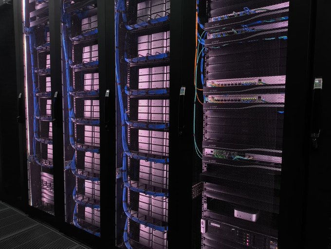 The image shows racks of stacked Mac Mini computers wired together as a computing cluster, with a Dell server visible at the bottom. The setup relates to the post by showing the physical "cluster" mentioned—a custom-built array of interconnected Mac Minis used to outperform a major Bitcoin fund, now with properly managed cables to address the previous fire hazard concern.