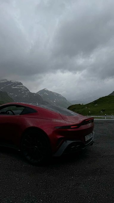 The rear view of a dark red sports car parked on asphalt against a dramatic backdrop of cloudy skies, mountains with patches of snow, and green hills. The license plate appears to read "AHB 2012".