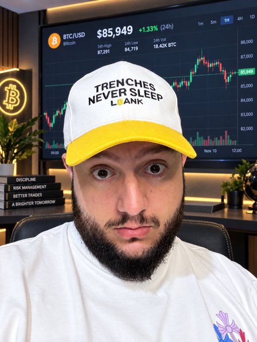A man with a beard wearing a white and yellow 'TRENCHES NEVER SLEEP' hat sits in front of a screen displaying a BTC/USD chart at $85,949.