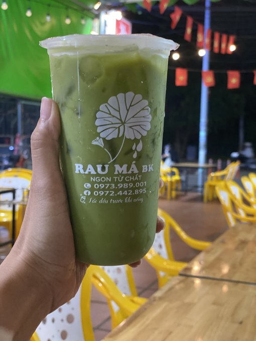 A hand holds a plastic cup of iced green tea from 'Rau Má BK' featuring a white lotus flower logo and Vietnamese text including a phone number.