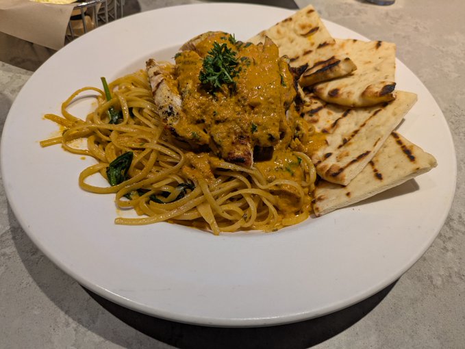 A white plate contains a meal of grilled chicken or fish topped with a yellow curry-like sauce and parsley, served alongside spaghetti pasta and pieces of grilled flatbread.