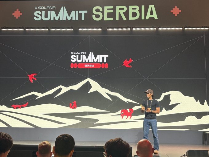 A man in a cap and jeans speaks on stage at the Solana Summit Serbia, with the event's logo and mountain graphics displayed on the large screen behind him.