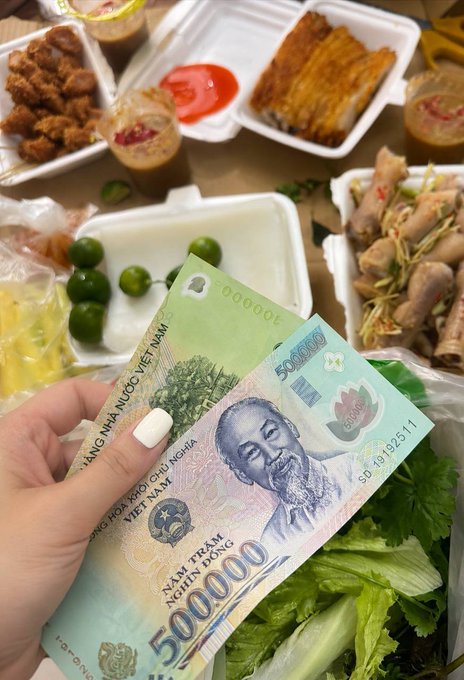 A hand with white nail polish holding a 500,000 Vietnamese Dong banknote over a spread of takeout food, including spring rolls, grilled meat, limes, and various dipping sauces.