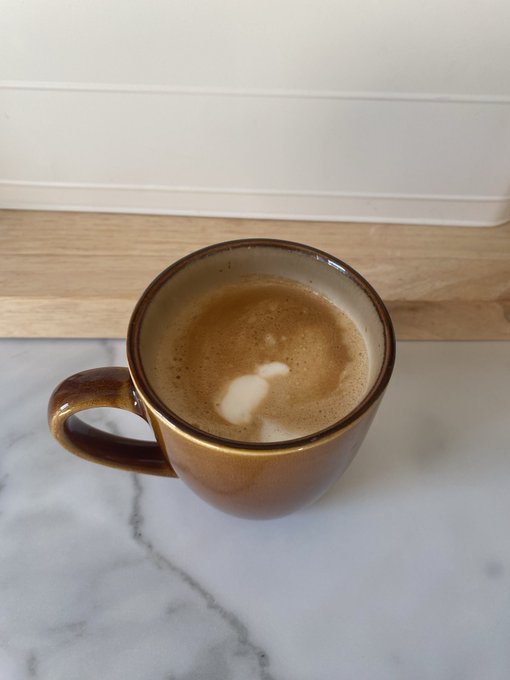 A brown ceramic mug filled with a foamy coffee drink, sitting on a white marble countertop against a light wood backsplash.