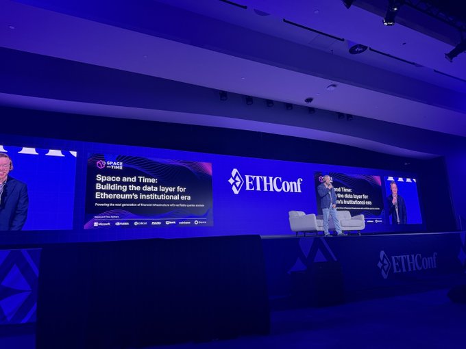 The image shows a large conference room with a stage set up for an event, featuring a speaker standing behind a podium. The stage has a blue backdrop that displays the text "Space & Time: Building the data layer for Ethereum". There are also other people present in the scene, possibly attendees or organizers of the event.