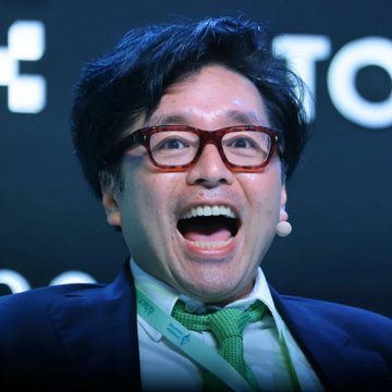 In the image, there's a man wearing glasses with red frames, dressed in a suit, and smiling. He has a green tie around his neck. The man seems to be making a funny or excited gesture. There are two numbers visible on the man, which might represent some sort of data related to the post he is sharing.