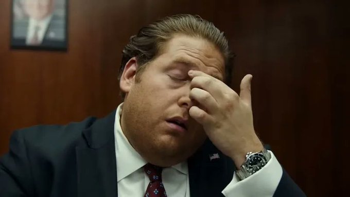 The image features a man wearing a suit and tie sitting at a desk, with his hand resting over his face as if he's deep in thought or experiencing some form of discomfort. The background includes a wooden wall and a picture hanging on the wall. There is also an advertisement for a product called "100 ETH" that was being sold for $2,050 on swapboard. However, the man appears to have missed this sale due to not plugging his bot into the API.