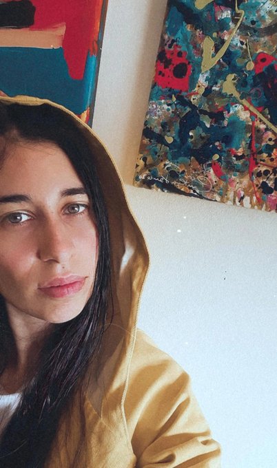 A young woman with long dark hair wears a yellow hoodie and gazes directly at the camera, standing in front of colorful abstract paintings in what appears to be an art studio, fitting the casual "GM" (Good Morning) greeting vibe.