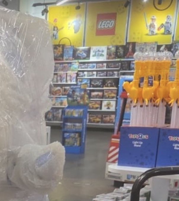 A photograph taken inside a toy store, likely Toys 'R' Us, showing shelves of games and toys with a LEGO branded wall, blue TOYS R US boxes in the foreground, and bubble-wrapped merchandise on the left.