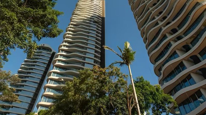 A low-angle photograph of modern, curved high-rise buildings with balconies against a clear blue sky, framed by green trees and a palm tree.