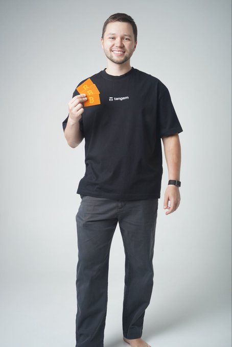 A smiling man in a black "tangem" t-shirt holds up an orange card with the Bitcoin logo. The image reflects his role in web3 at Tangem, visually tying his personal brand to the crypto and self-custody work he describes.